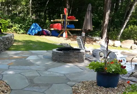 Stone fire pit patio beside a wooded lakeside home