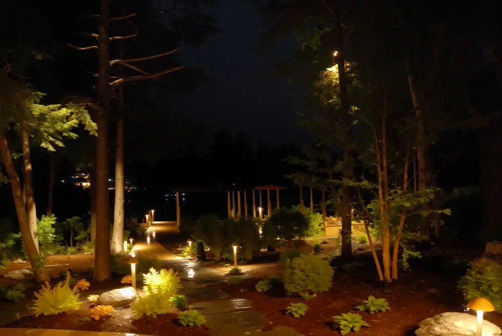 Low-voltage path lights illuminating a wooded garden walkway beside the lake at night